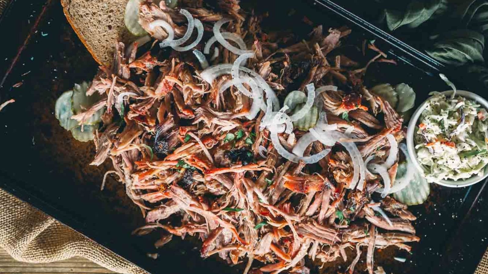 Shredded smoked picnic shoulder topped with sliced onions, served with pickles, bread, and a side of coleslaw on a dark tray.