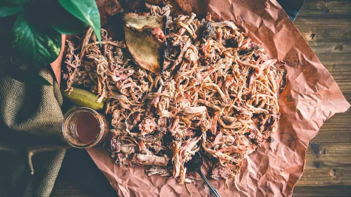 Pulled pork served on brown butcher paper with a bone, barbecue sauce in a jar, a pickle slice, and a fork on a wooden surface.