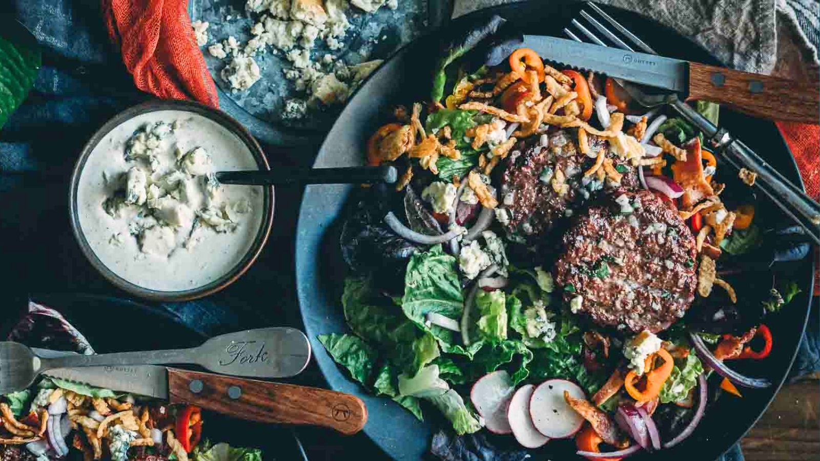 A plate of fresh salad topped with two grilled burger patties, sliced radishes, peppers, crispy onions, and crumbled cheese, with a bowl of creamy dressing on the sideโperfect for Father's Day recipes.