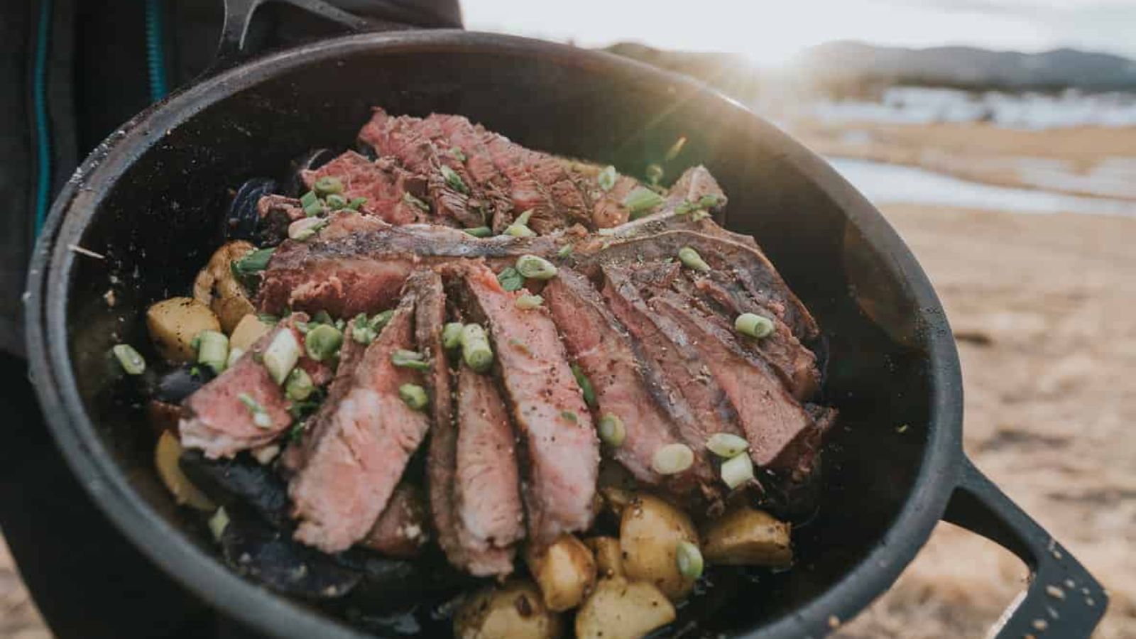 A cast iron skillet filled with sliced steak, diced potatoes, and chopped green onions is held outdoors with a scenic background.