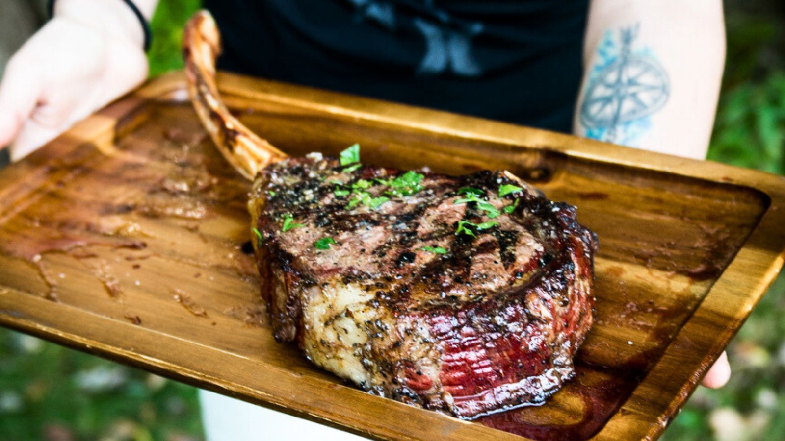 A person holds a wooden tray with a cooked tomahawk steak garnished with chopped herbs.