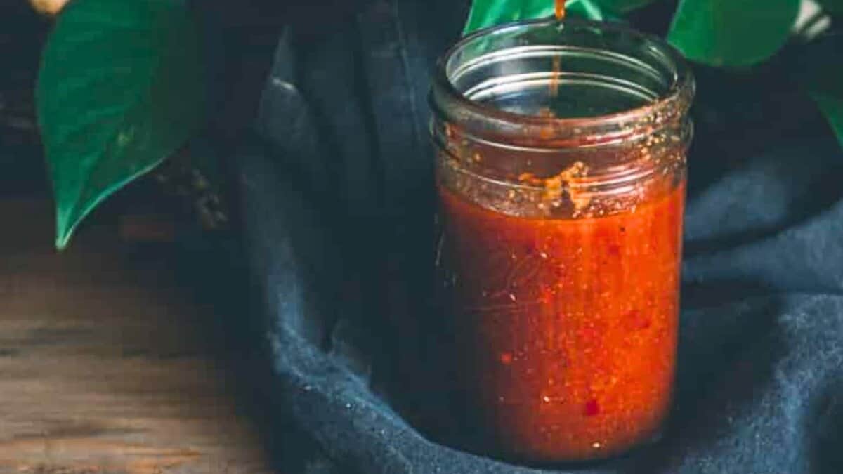 A glass jar filled with red chili sauce sits on a dark cloth, with green leaves partially visible in the background.