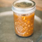 A glass mason jar filled with chunky brown sauce or chutney, sealed with a metal lid, sits on a gray surface.