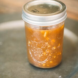 A glass mason jar filled with chunky brown sauce or chutney, sealed with a metal lid, sits on a gray surface.