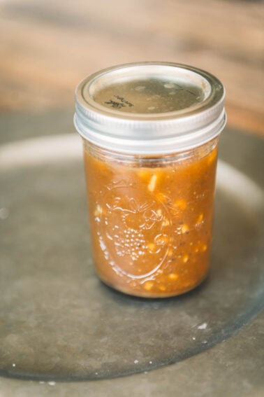 A glass mason jar filled with chunky brown sauce or chutney, sealed with a metal lid, sits on a gray surface.
