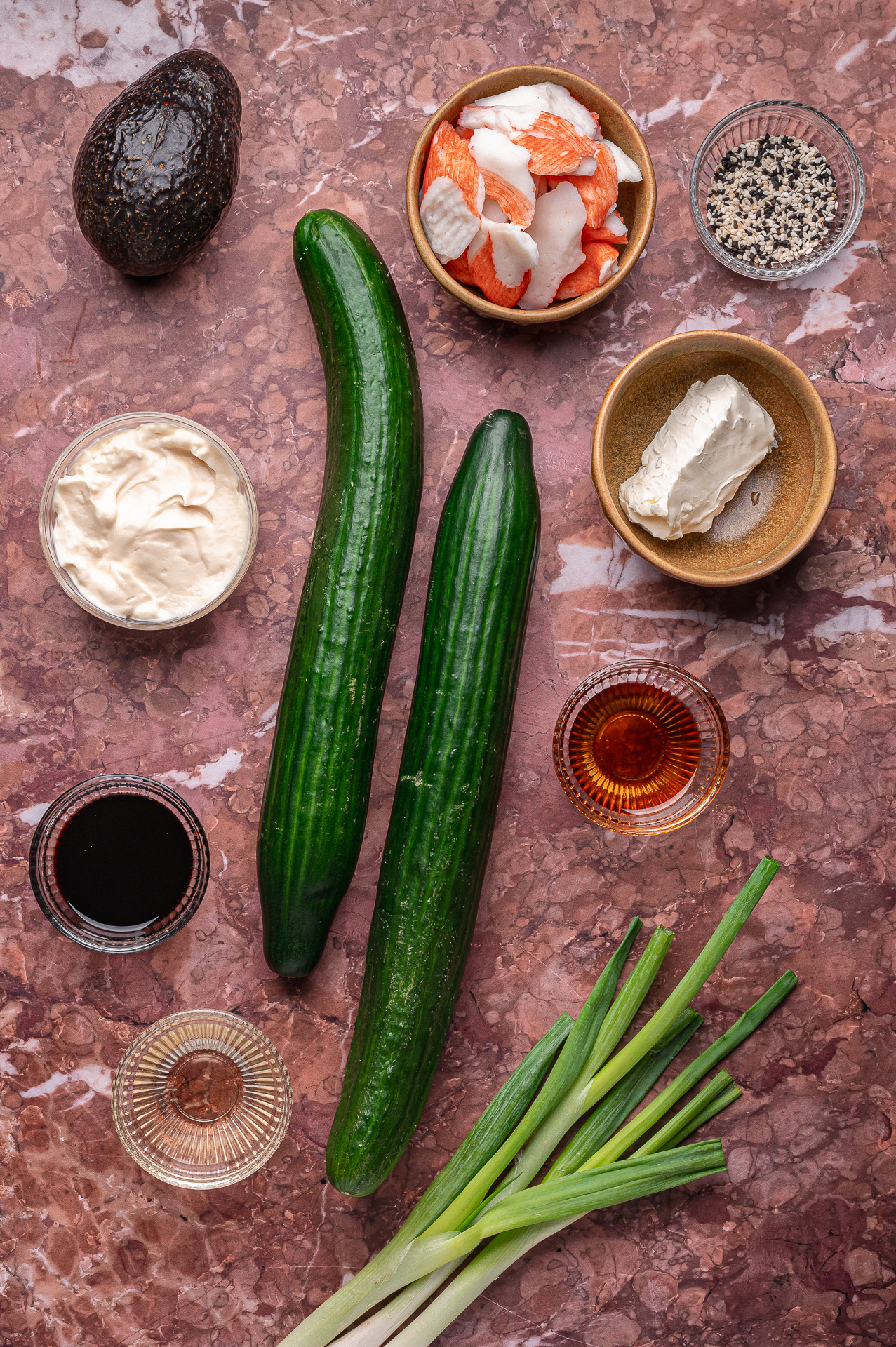 A flat lay of sushi ingredients including two cucumbers, imitation crab, avocado, cream cheese, mayonnaise, green onions, soy sauce, sesame seeds, and rice vinegar on a marbled surface.