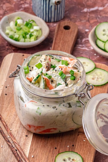 A glass jar filled with creamy salad containing sliced cucumber, carrots, and green onions, placed on a wooden board with extra vegetables and sesame seeds nearby.