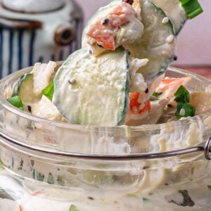 A close-up of chopsticks lifting creamy cucumber salad with crab and sesame from a glass jar; a ceramic container is in the background.