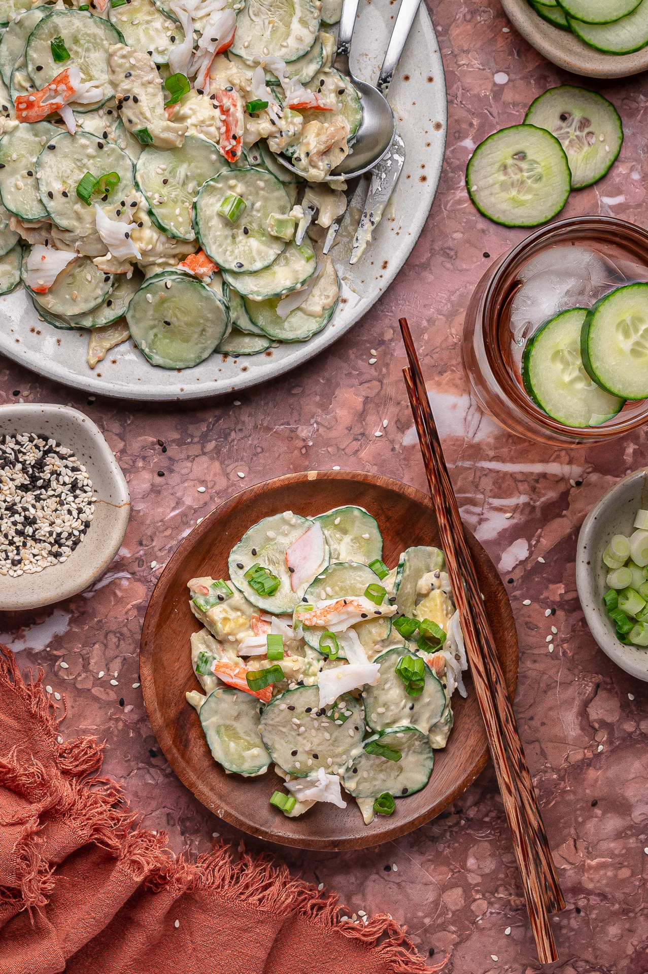 Plate and bowl of creamy cucumber salad with crab sticks, topped with sesame seeds and green onions; chopsticks, cucumber slices, seasoning, and a glass of ice water nearby on a marble surface.