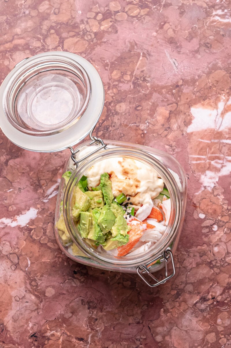 A glass jar with an open lid containing avocado, imitation crab sticks, mayonnaise, onions, and sesame seeds, on a marbled pink surface.