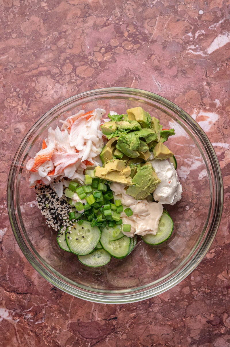 A glass bowl containing sliced cucumber, imitation crab, avocado chunks, cream cheese, mayonnaise, sesame seeds, and chopped green onions on a marbled surface.