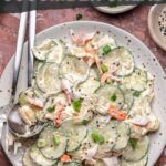 A plate of California roll cucumber salad with imitation crab, cucumber slices, and creamy dressing, garnished with green onions, served with a fork and spoon.