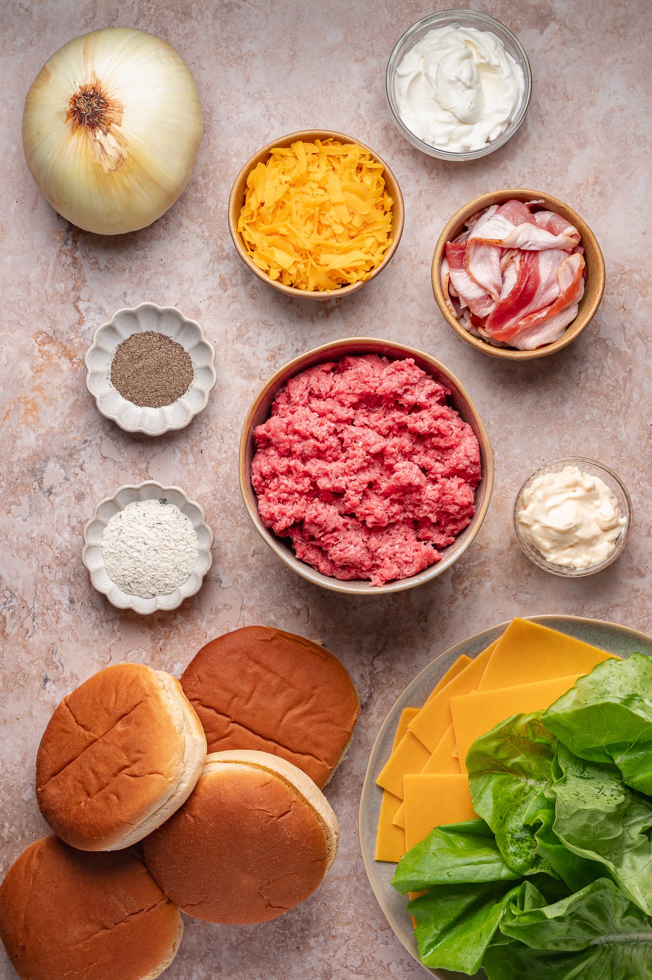 Ingredients for burgers arranged on a counter, including ground beef, shredded cheddar, bacon, onion, lettuce, sliced cheese, burger buns, mayonnaise, sour cream, and seasonings.