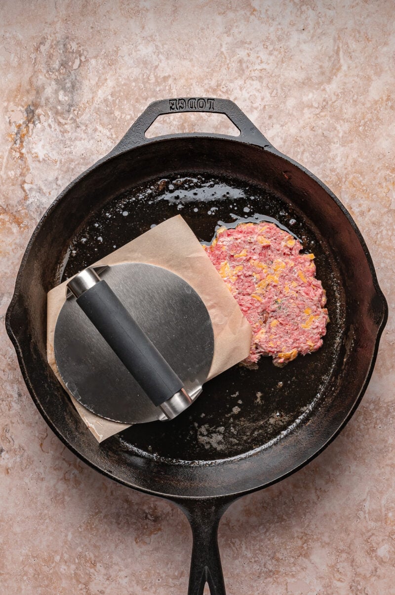 A raw burger patty is being pressed with a metal burger press and parchment paper in a cast iron skillet.