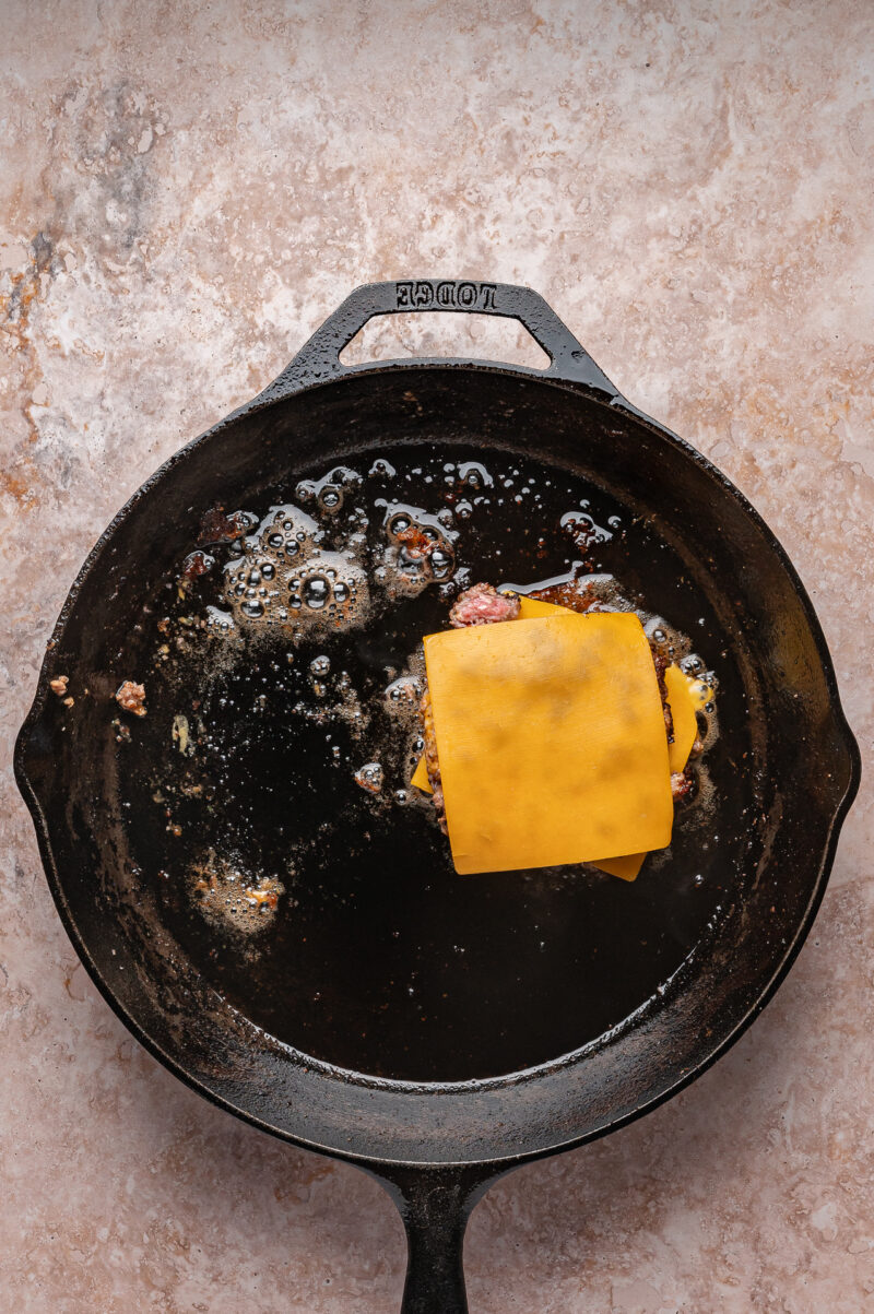 A cast iron skillet with grease and remnants of cooked food, featuring a partially melted slice of cheddar cheese on top of a small piece of meat.