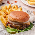 A cheeseburger with lettuce, onions, and a toasted bun served with crinkle-cut fries on newspaper-style paper.