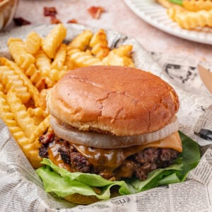 A cheeseburger with lettuce, onions, and a toasted bun served with crinkle-cut fries on newspaper-style paper.