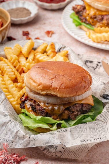 A cheeseburger with lettuce, onions, and a toasted bun served with crinkle-cut fries on newspaper-style paper.