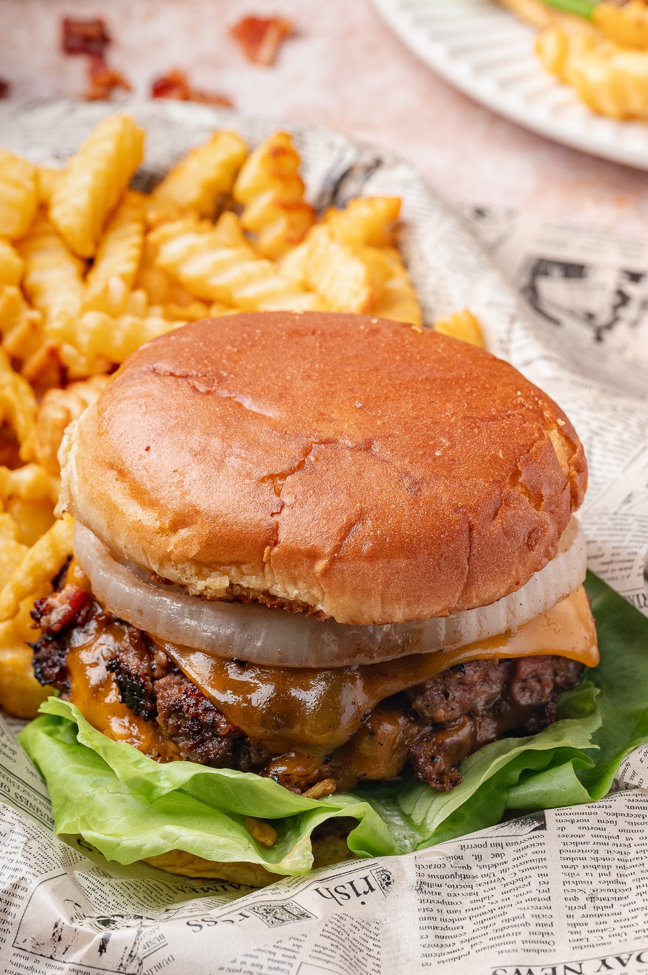 A cheeseburger with lettuce, onion, and cheddar cheese on a bun, served with crinkle-cut fries on newspaper-style paper.