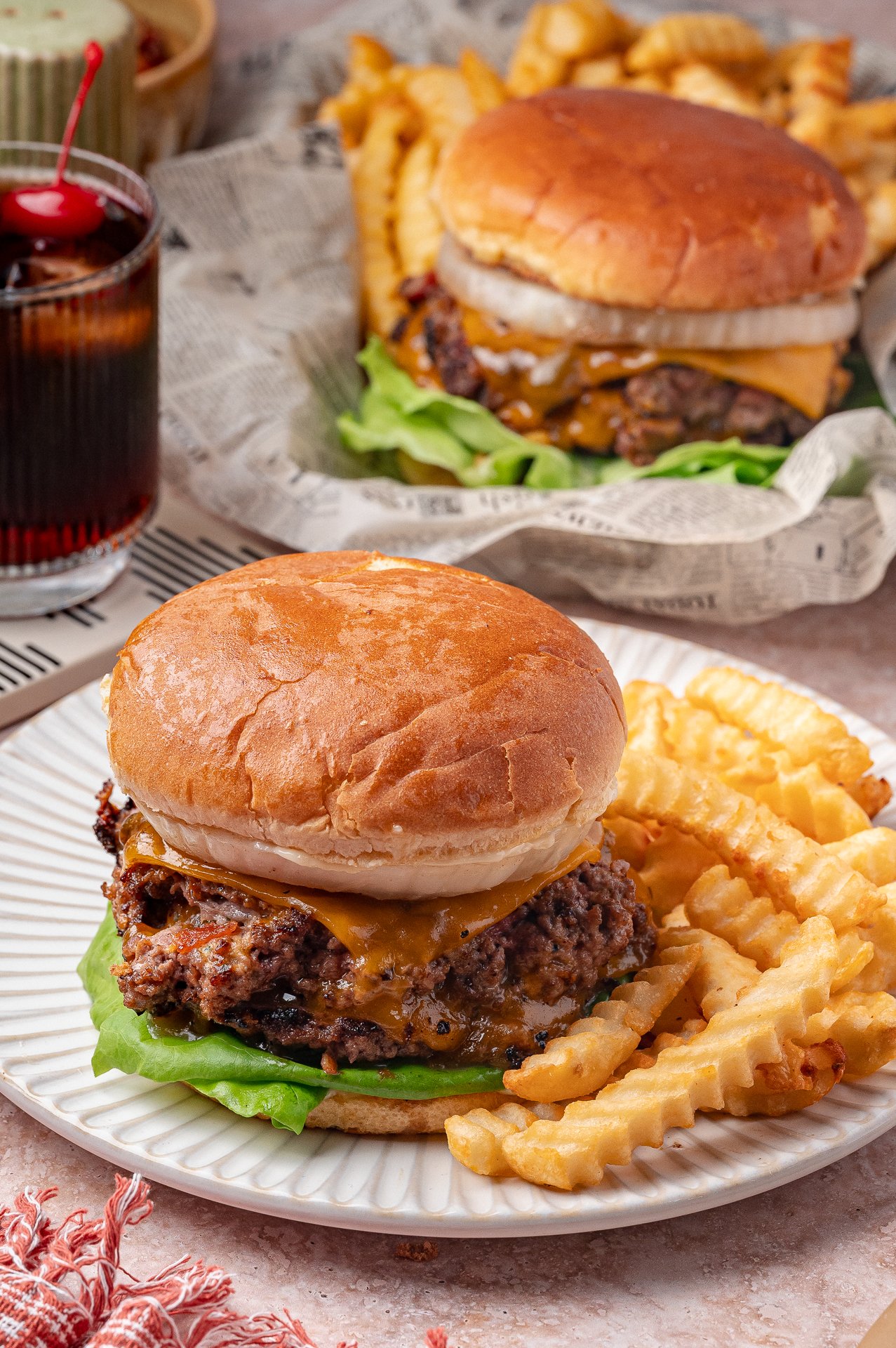 A cheeseburger with lettuce and onion on a bun is served with crinkle-cut fries on a white plate; another similar meal and a drink are in the background.