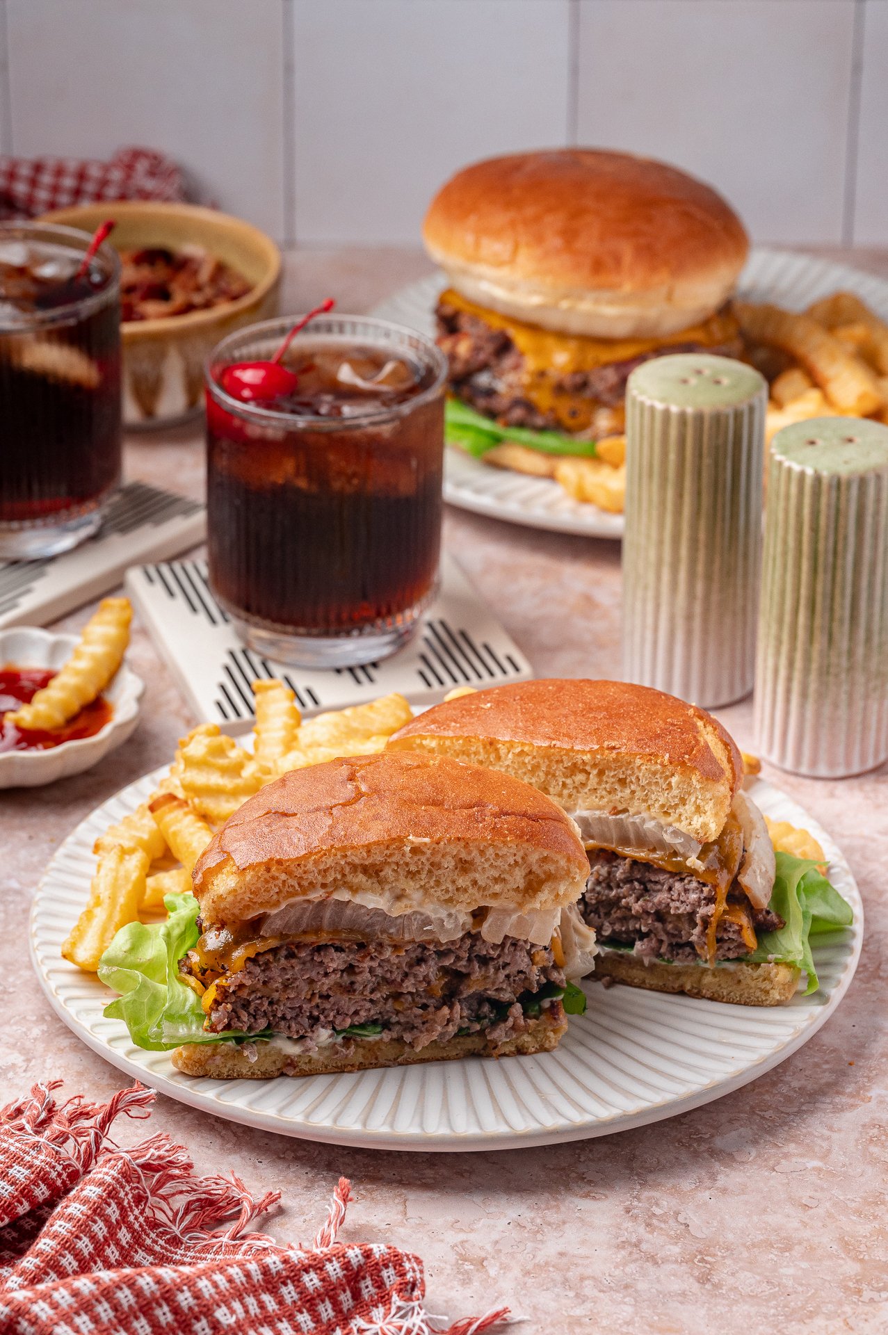 A plate with a halved cheeseburger, lettuce, fries, and a drink on a table; another burger, fries, and drinks are in the background.