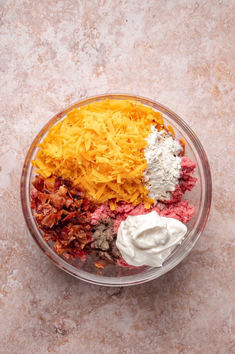 A glass bowl containing shredded cheddar cheese, flour, ground beef, cooked bacon pieces, black pepper, and a dollop of sour cream, on a light stone surface.