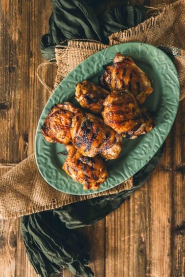 Grilled chicken thighs arranged on a green oval plate, set on a wooden table with burlap and dark green fabric accents.