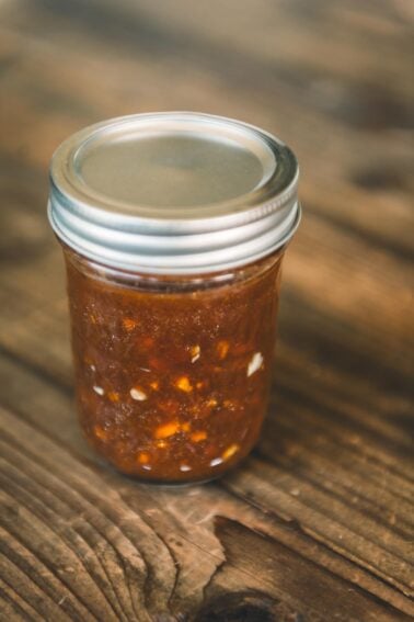 A small glass jar with a metal lid filled with chunky brown jam or preserve, sitting on a wooden surface.