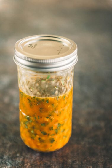 A glass jar with a metal lid, filled with an orange sauce containing visible herbs and spices, sits on a textured surface.