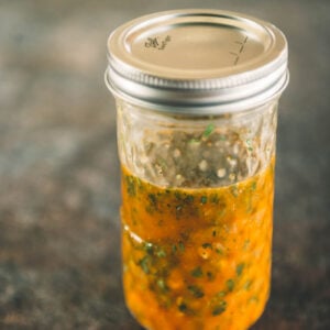A glass jar with a metal lid containing orange-colored sauce with visible herbs, placed on a textured brown surface.