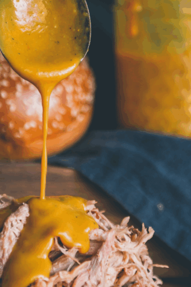 Shredded meat on a sandwich bun with yellow sauce being poured over it; more buns and a jar of sauce are in the background.