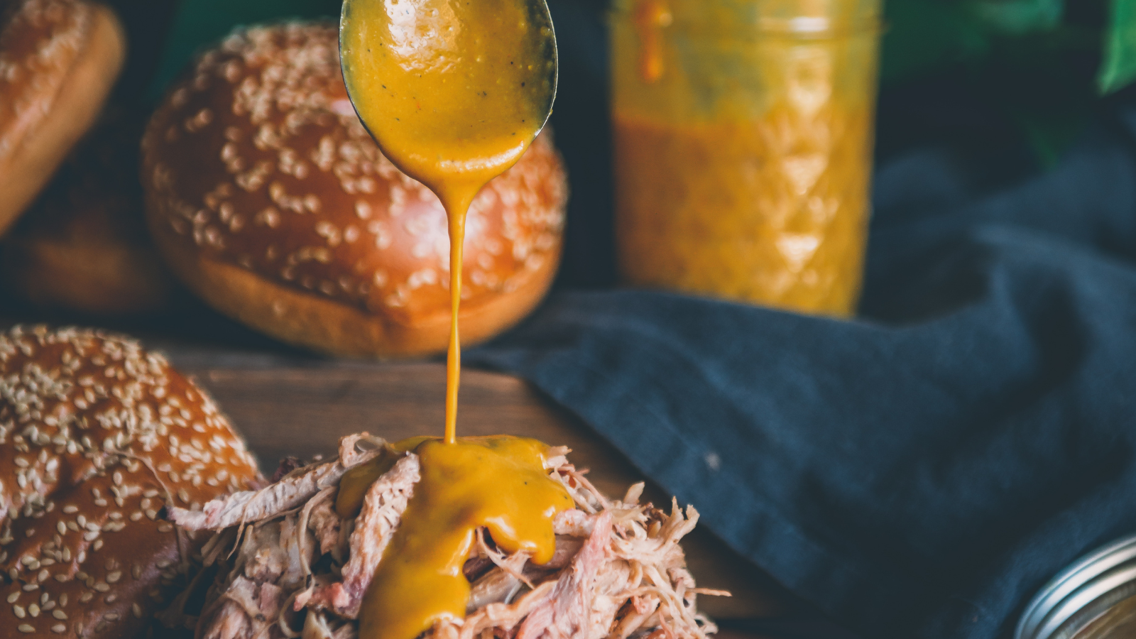 Shredded meat on a sandwich bun with yellow sauce being poured over it; more buns and a jar of sauce are in the background.