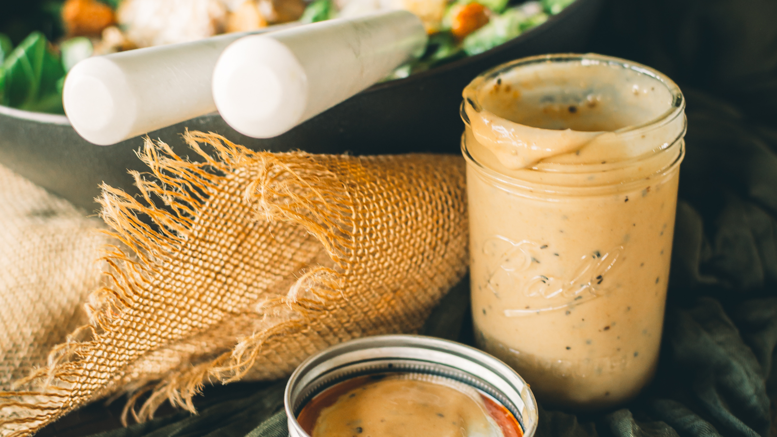 A glass jar filled with creamy salad dressing, with its lid resting beside it, sits next to a kitchen towel and a salad bowl.