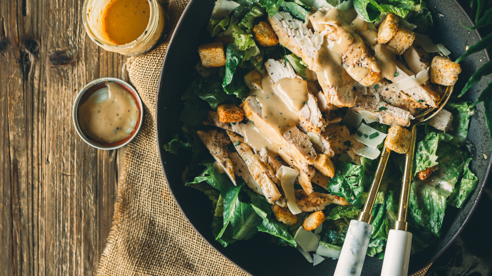 A plate of Caesar salad with grilled chicken, croutons, shaved cheese, and dressing, next to a jar and small bowl of extra dressing on a rustic wooden table.