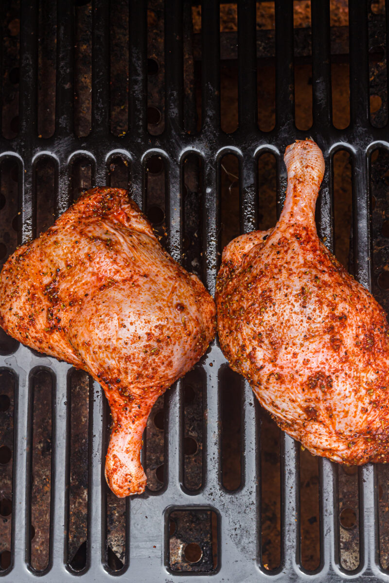 Two seasoned duck leg quarters placed side by side on a grill grate, ready to be cooked.