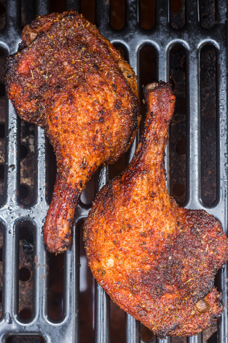 Two seasoned, smoked duck leg quarters cooking on a metal grill.