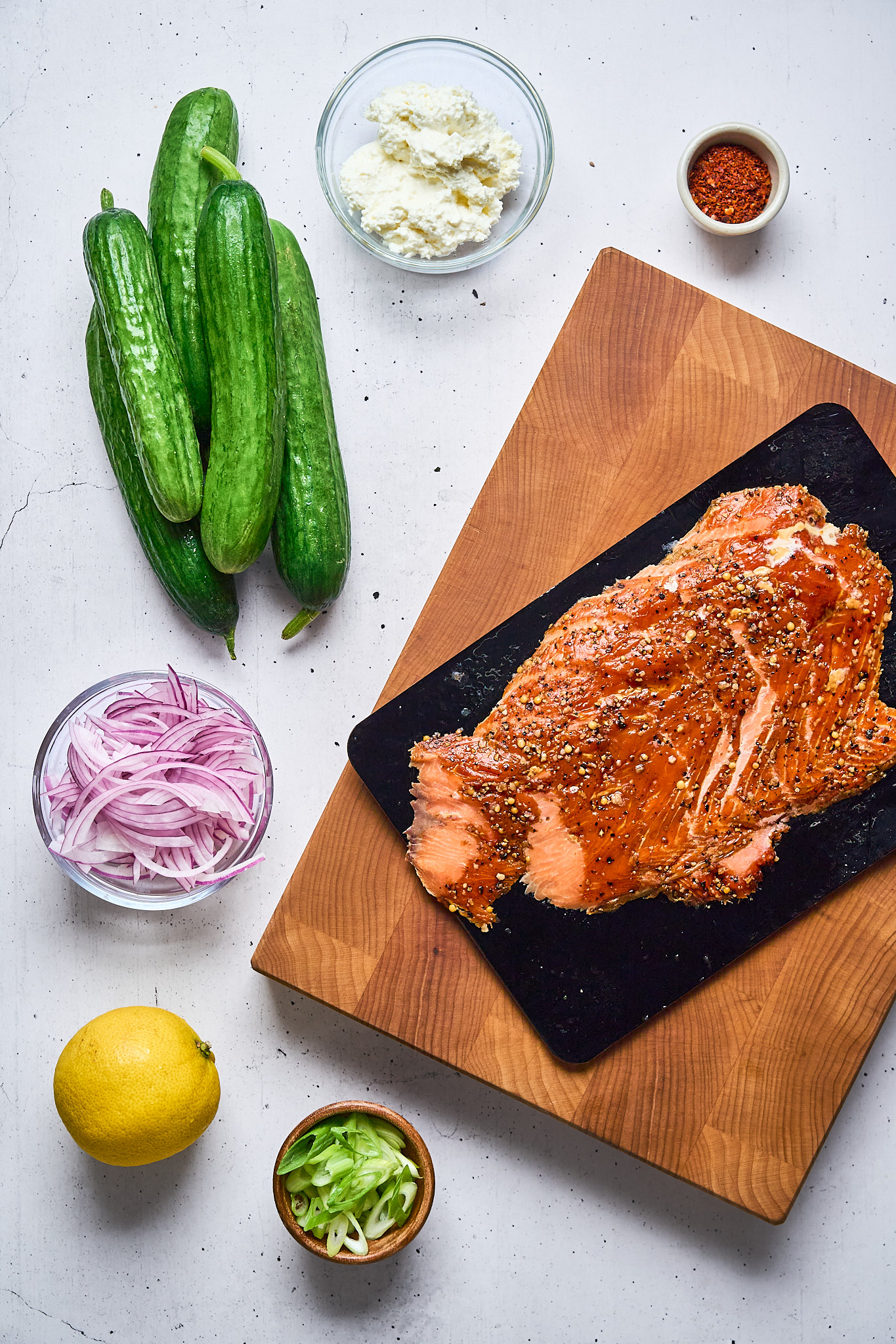 Sliced smoked salmon on a cutting board, surrounded by cucumbers, red onion, lemon, scallions, crumbled cheese, and a small bowl of red pepper flakes on a white surface.