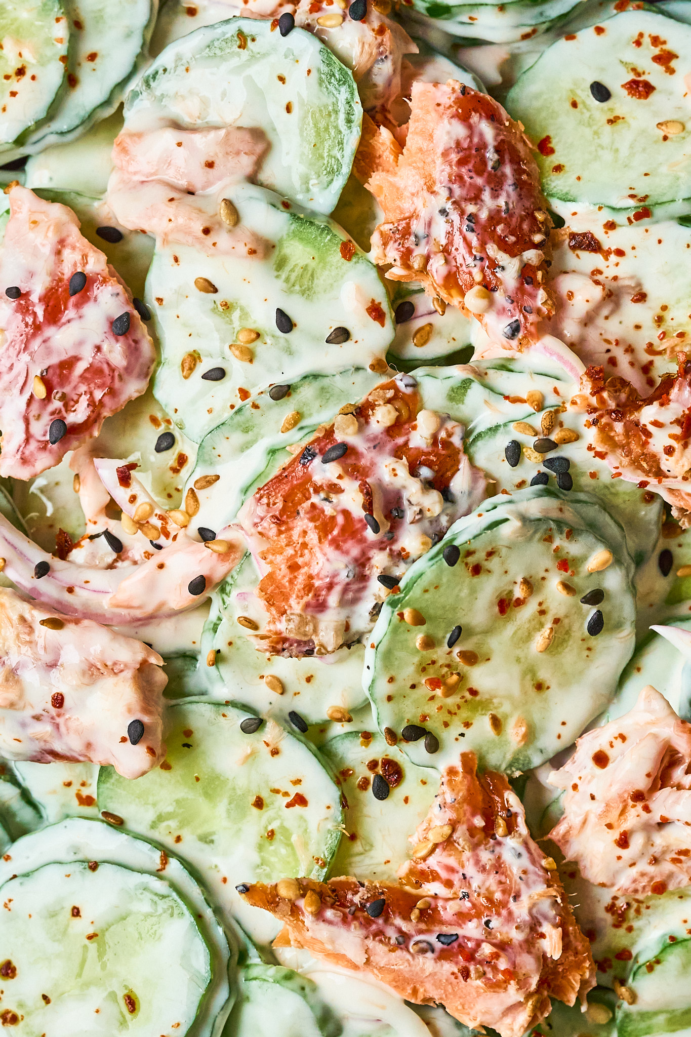 Close-up of Philly roll cucumber salad, sliced cucumbers and pieces of salmon mixed in a creamy dressing, topped with black sesame seeds and red pepper flakes.