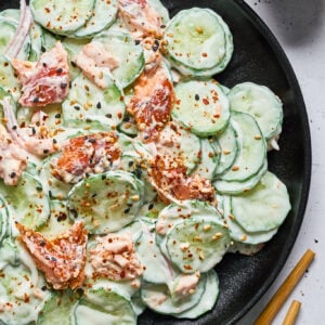 A black plate with cucumber salad dressed in a creamy sauce, topped with smoked salmon and sprinkled with sesame seeds and chili flakes. A small bowl of seeds and chopsticks are nearby.