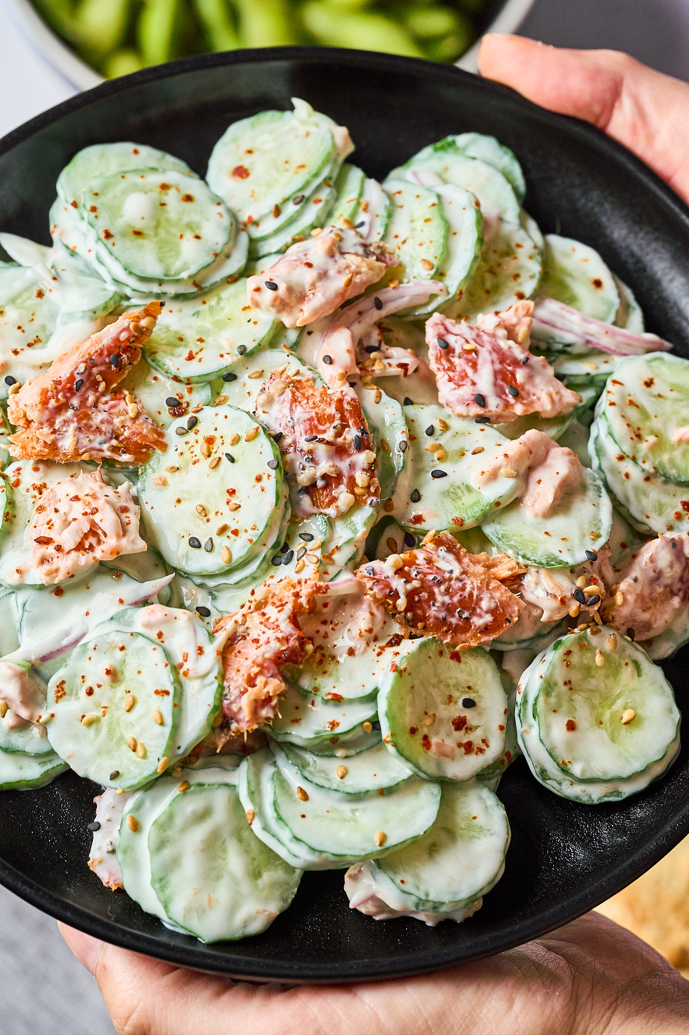 A black plate with sliced cucumbers mixed with creamy dressing, shredded smoked salmon, and sprinkled with red pepper flakes.