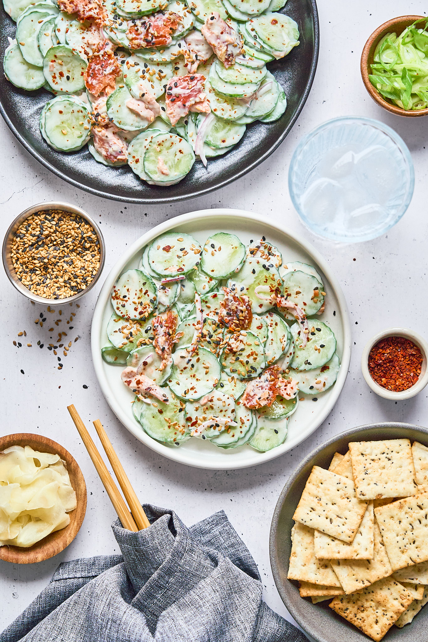 A white plate of cucumber salad with sliced cucumbers, smoked salmon, dressing, and red pepper flakes, surrounded by crackers, lettuce, seasoning, a glass of water, and chopsticks.