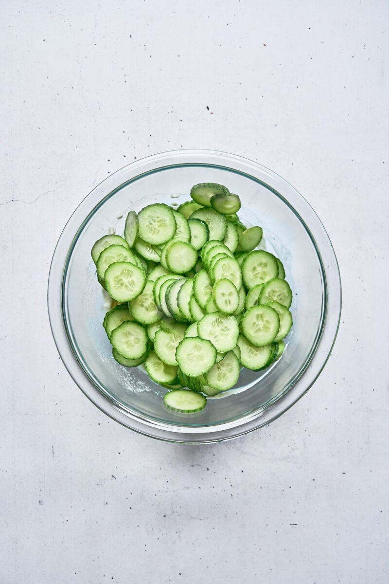 A glass bowl filled with thinly sliced cucumbers sits on a white surface.