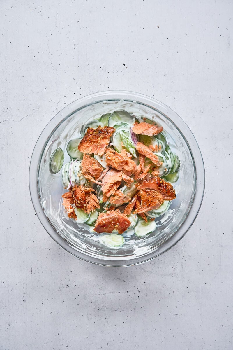 A glass bowl containing a salad made of sliced cucumbers, creamy dressing, and flaked pieces of smoked salmon.