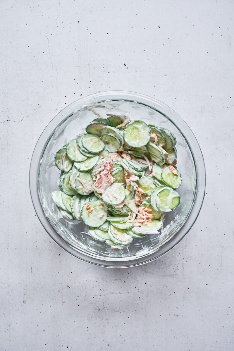 A glass bowl filled with sliced cucumbers and a creamy dressing, placed on a light-colored surface.