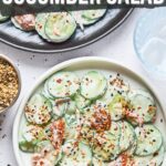 A bowl of cucumber salad topped with shredded smoked salmon and sprinkled with sesame seeds and seasonings, next to a small bowl of spices.