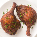 Two seasoned and cooked duck legs garnished with chopped parsley on a white plate.