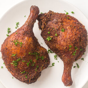 Two seasoned and cooked duck legs garnished with chopped parsley on a white plate.