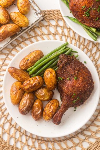 A white plate with roasted baby potatoes, green beans, and a smoked duck leg, placed on a woven placemat. Another plate and a tray with potatoes are partially visible.