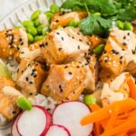 A bowl with rice, sesame-crusted salmon, sliced radishes, shredded carrots, edamame, fresh cilantro, and a drizzle of creamy sauce.