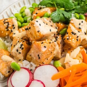 A bowl with rice, sesame-crusted salmon, sliced radishes, shredded carrots, edamame, fresh cilantro, and a drizzle of creamy sauce.